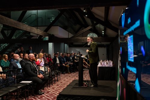 A large group of award show attendees sit in a slightly darkened function room while the Australian Web Awards MC speaks on stage